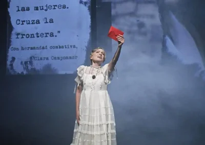 Escena de la ópera Hildegart con una mujer alzando una carta roja, sobre fondo de proyección con cita de Clara Campoamor